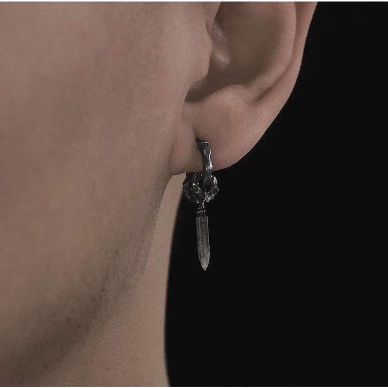 May include: A single silver earring with a dagger pendant. The earring is attached to the ear with a small hoop. The dagger is a long, thin blade with a pointed tip. The earring is worn on the ear.