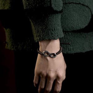 May include: A dark brown bracelet with a silver infinity symbol charm. The bracelet is made of woven cord and is worn on a wrist. The background is dark, with a green sweater sleeve visible.