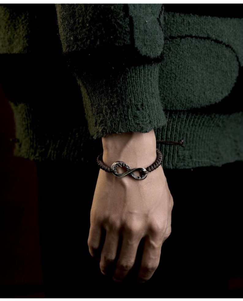 May include: A dark brown bracelet with a silver infinity symbol charm. The bracelet is made of woven cord and is worn on a wrist. The background is dark, with a green sweater sleeve visible.