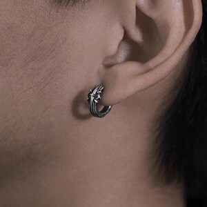 May include: A small silver hoop earring featuring an intricate, raised cross design on the outer surface. The metal has a dark, oxidized finish within the recessed areas, highlighting the detailed texture of the cross motif on the circular band.
