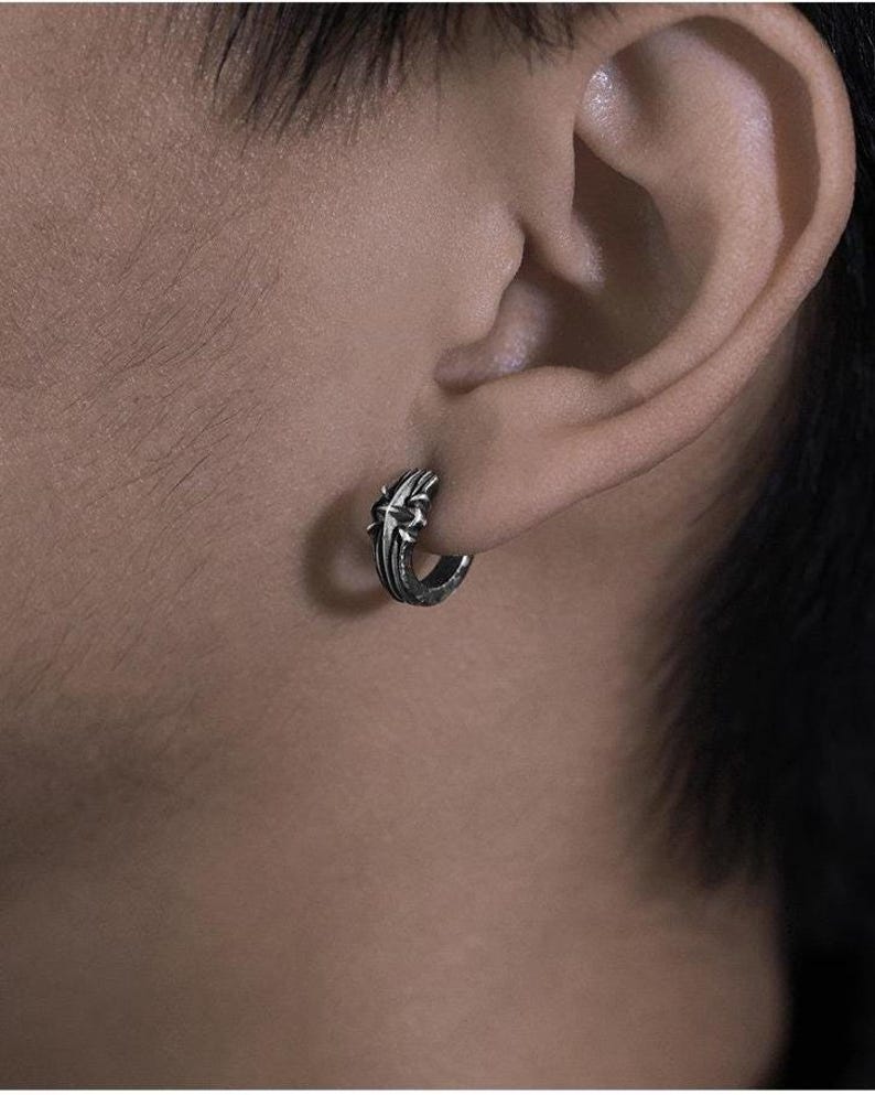 May include: A small silver hoop earring featuring an intricate, raised cross design on the outer surface. The metal has a dark, oxidized finish within the recessed areas, highlighting the detailed texture of the cross motif on the circular band.