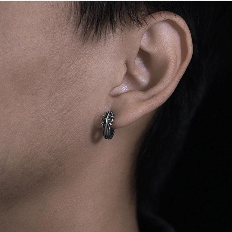 May include: A small, circular hoop earring made of oxidized silver-toned metal. The earring features a raised, textured cross design on its outer surface, creating a detailed, dark, and metallic appearance that sits securely on the earlobe.
