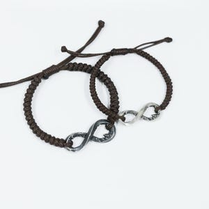 May include: Two brown braided cord bracelets with silver infinity symbol charms. The bracelets have adjustable sliding knots. The infinity charms have a textured, slightly distressed appearance, and are connected to the cord.