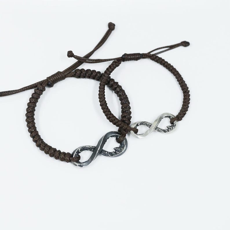 May include: Two brown braided cord bracelets with silver infinity symbol charms. The bracelets have adjustable sliding knots. The infinity charms have a textured, slightly distressed appearance, and are connected to the cord.