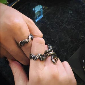Pu&ograve; includere: Primo piano di tre anelli argentati. Due anelli presentano un design attorcigliato, mentre il terzo ha la forma di un serpente con dettagli verdi e dorati. Gli anelli sono indossati alle dita.