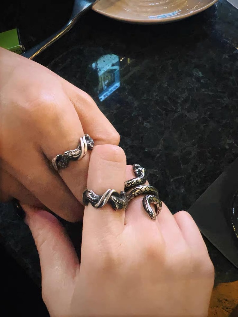 Pu&ograve; includere: Primo piano di tre anelli argentati. Due anelli presentano un design attorcigliato, mentre il terzo ha la forma di un serpente con dettagli verdi e dorati. Gli anelli sono indossati alle dita.