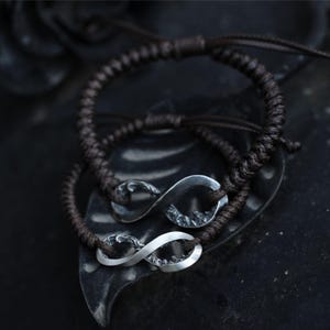 May include: Two brown braided cord bracelets with silver infinity symbols. The bracelets are displayed on a dark, textured surface. The infinity symbols have a detailed, textured design.