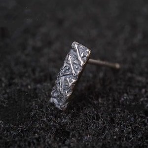 Peut inclure: Boucle d'oreille unique en argent avec un design rectangulaire. La boucle d'oreille présente une surface texturée avec des motifs complexes et une tige pour l'insertion. L'arrière-plan est sombre et texturé.