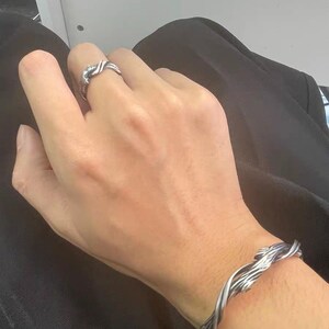Pu&ograve; includere: Un set di anello e bracciale argentati. L'anello presenta un design intrecciato e il bracciale ha un motivo simile. I gioielli sembrano essere in metallo e sono indossati sulla mano e sul polso di una persona.