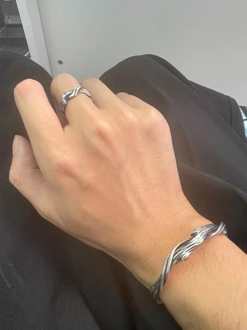 Pu&ograve; includere: Un set di anello e bracciale argentati. L'anello presenta un design intrecciato e il bracciale ha un motivo simile. I gioielli sembrano essere in metallo e sono indossati sulla mano e sul polso di una persona.