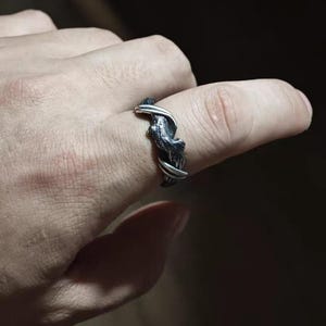 Pu&ograve; includere: Un anello d'argento con una fascia strutturata grigio scuro e un design organico attorcigliato. L'anello &egrave; indossato su un dito, mostrando i suoi dettagli intricati e le trame contrastanti. Il design dell'anello assomiglia a rami intrecciati.
