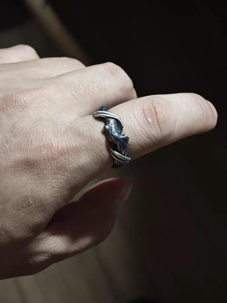 Pu&ograve; includere: Un anello d'argento con una fascia strutturata grigio scuro e un design organico attorcigliato. L'anello &egrave; indossato su un dito, mostrando i suoi dettagli intricati e le trame contrastanti. Il design dell'anello assomiglia a rami intrecciati.