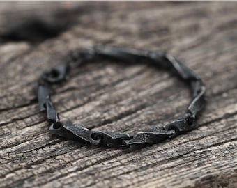 May include: A dark gray metal bracelet with a textured, link design. The bracelet is made of individual, rectangular links with a slightly twisted appearance. The bracelet is displayed on a weathered wooden surface.