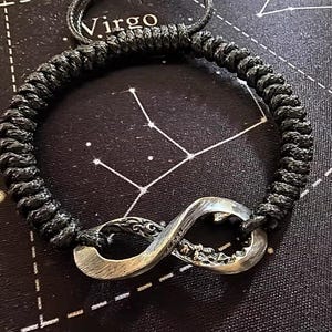 May include: Black braided cord bracelet with a silver infinity symbol charm. The charm has intricate detailing. The word "Virgo" is visible in the background. The bracelet is a stylish accessory.