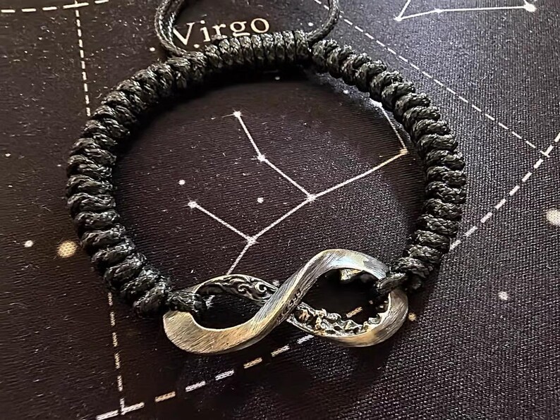 May include: Black braided cord bracelet with a silver infinity symbol charm. The charm has intricate detailing. The word "Virgo" is visible in the background. The bracelet is a stylish accessory.