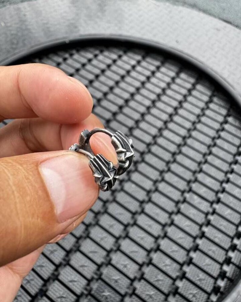 May include: A small silver hoop earring featuring a textured, multi-faceted design with pointed, star-like details along the outer edge. The metal has a darkened, oxidized finish in the recessed areas, highlighting the intricate, geometric relief pattern.