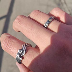 Pu&ograve; includere: Due anelli d'argento sono mostrati su dita. Un anello ha un design intrecciato con un accento dorato, mentre l'altro presenta un motivo fluido e strutturato. Gli anelli sono su uno sfondo sfocato.