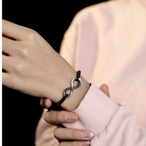 May include: Black braided cord bracelet featuring a silver infinity symbol. The infinity symbol has a textured design. The bracelet is worn on a wrist, against a pink long-sleeved shirt.
