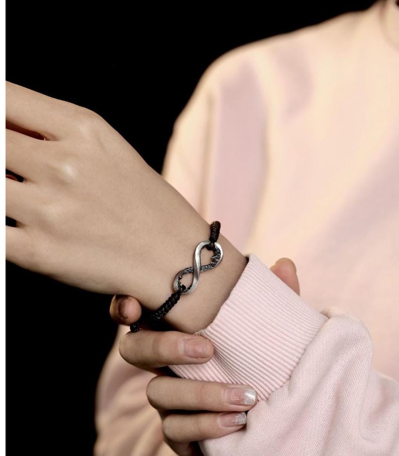 May include: Black braided cord bracelet featuring a silver infinity symbol. The infinity symbol has a textured design. The bracelet is worn on a wrist, against a pink long-sleeved shirt.