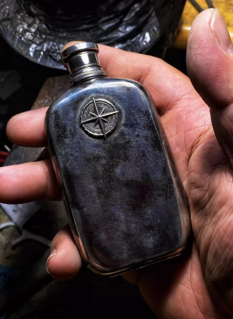 Pu&ograve; includere: Una fiaschetta d'argento con un design a bussola sul davanti. La fiaschetta &egrave; tenuta in una mano.