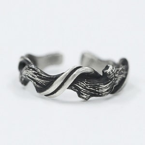 Pu&ograve; includere: Anello color argento con un design strutturato e organico. L'anello presenta un motivo a spirale, simile a un'onda, con fascia divisa. Il metallo ha una finitura leggermente scurita e antica, su sfondo bianco.
