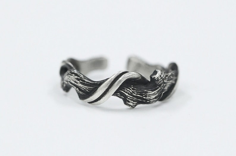 Pu&ograve; includere: Anello color argento con un design strutturato e organico. L'anello presenta un motivo a spirale, simile a un'onda, con fascia divisa. Il metallo ha una finitura leggermente scurita e antica, su sfondo bianco.