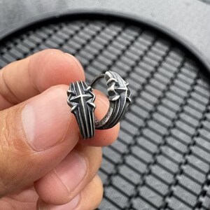 May include: A silver-toned hoop earring featuring a raised, textured cross design on the outer surface. The metal has an oxidized, darkened finish in the recessed areas, highlighting the detailed, angular structure of the cross motif.