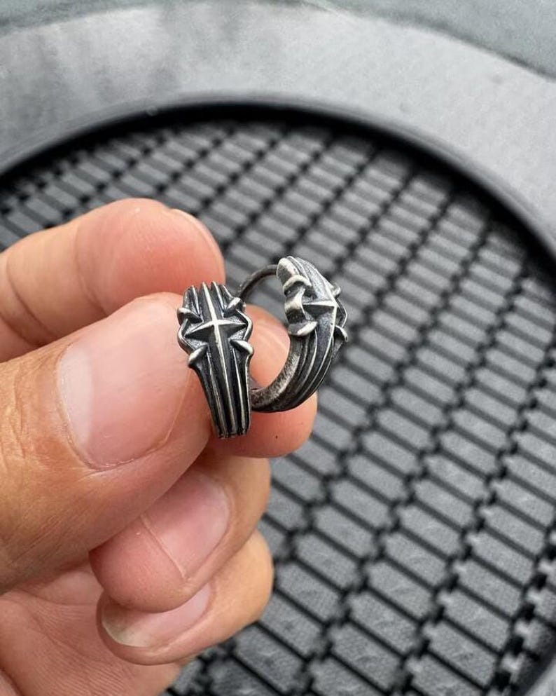 May include: A silver-toned hoop earring featuring a raised, textured cross design on the outer surface. The metal has an oxidized, darkened finish in the recessed areas, highlighting the detailed, angular structure of the cross motif.