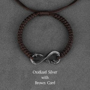 May include: A brown cord bracelet with an oxidized silver infinity symbol. The bracelet is knotted and has a sliding knot closure. The text "Oxidized Silver with Brown Cord" is below the bracelet.