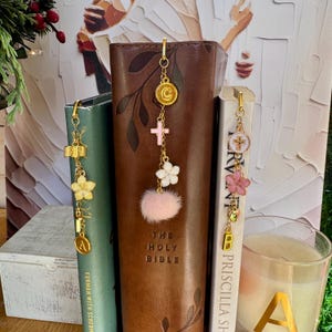 Puede incluir: Marcadores dorados con colgantes que se muestran junto a libros. Los colgantes incluyen cruces, flores y un pompón rosa. Un libro está etiquetado como "The Holy Bible". Los marcadores están sujetos a la parte superior de los libros.