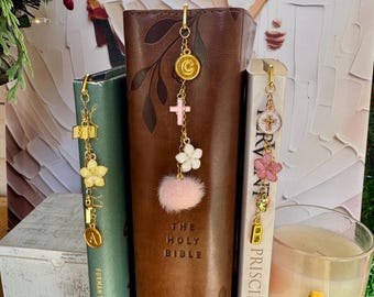 May include: Gold-toned bookmarks with dangling charms are shown next to books. The charms include crosses, flowers, and a pink pom-pom. One book is labeled "The Holy Bible". The bookmarks are attached to the top of the books.