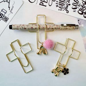 May include: Gold-colored metal cross-shaped paper clips with decorative charms. One clip holds a pen with a floral pattern. Charms include a pink pom-pom, and gold-colored letters. Other paper clips are plain.