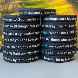May include: A collection of black silicone wristbands, each inscribed with a different Bible verse in white and colored text. Verses include "Encourage one another" and "I can do all things." The bands are stacked on a light surface.