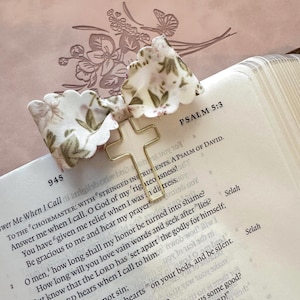 May include: A gold-colored cross-shaped paper clip with a floral fabric bow attached, resting on the open pages of a book. The text "PSALM 5:3" is visible, along with other biblical verses. The bow features a white background with green and brown floral patterns.