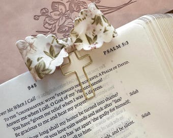 May include: A gold-colored cross-shaped paper clip with a floral fabric bow attached, resting on the open pages of a book. The text "PSALM 5:3" is visible, along with other biblical verses. The bow features a white background with green and brown floral patterns.