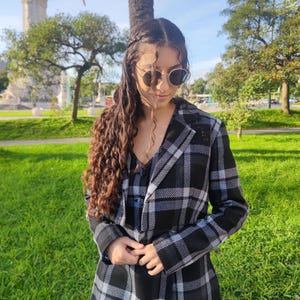 May include: A woman wearing a black and white plaid jacket with a black and white patterned top underneath. She is wearing round sunglasses and has long, curly brown hair.