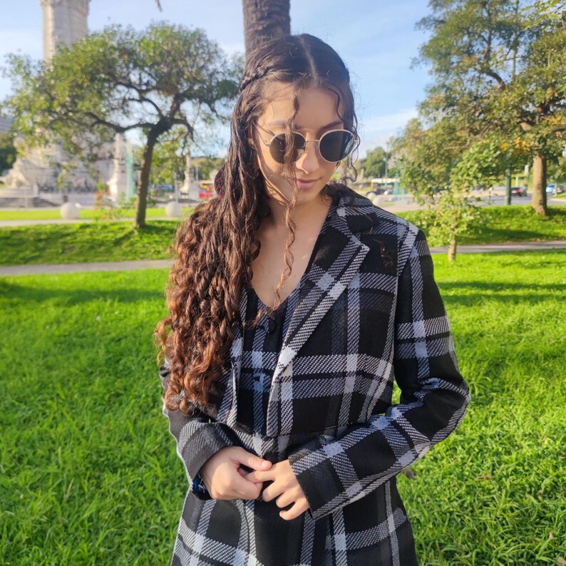 May include: A woman wearing a black and white plaid jacket with a black and white patterned top underneath. She is wearing round sunglasses and has long, curly brown hair.