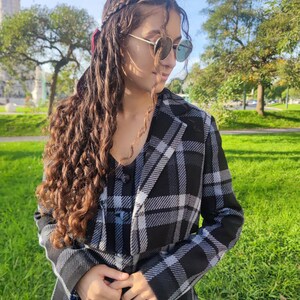 May include: A woman wearing a black and white plaid jacket with a long, curly brown hairstyle. She is wearing sunglasses and looking down.