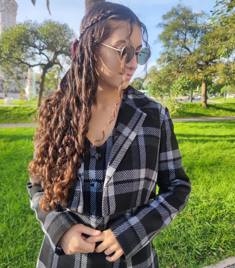 May include: A woman wearing a black and white plaid jacket with a long, curly brown hairstyle. She is wearing sunglasses and looking down.