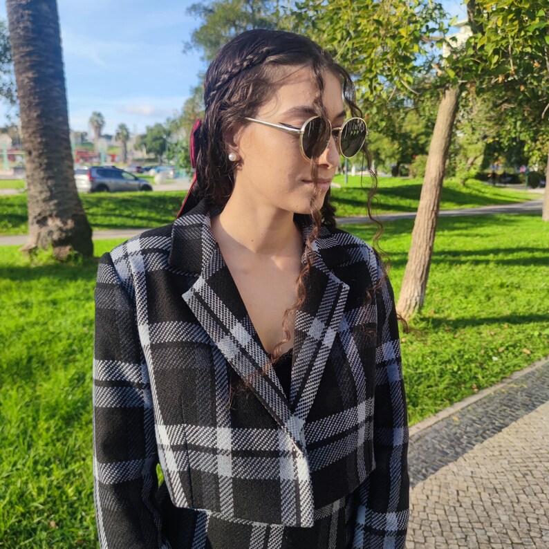 May include: A woman wearing a black and white plaid cropped jacket with a red ribbon in her hair. She is wearing sunglasses and has her hair pulled back in a braid.