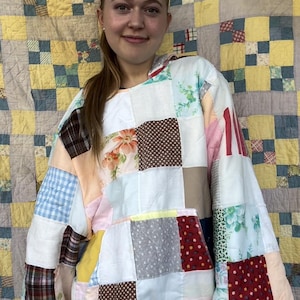 May include: A white hooded sweatshirt made from patchwork fabric with different patterns and colors. The sweatshirt has a large pocket on the front and is worn over a pair of blue jeans.