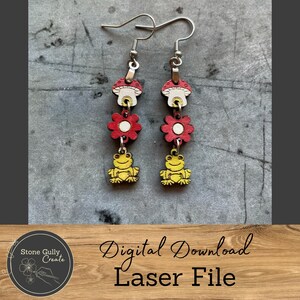 May include: A pair of dangle earrings featuring a mushroom, flower, and frog design. The earrings have silver-colored hooks and are made of wood. The mushroom is red and white, the flower is red, and the frog is yellow.
