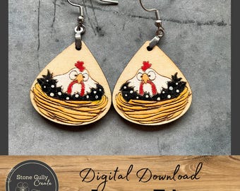 May include: A pair of laser cut wooden earrings featuring a cartoon chicken in a nest. The chicken is white with black and white spots and red accents. The nest is brown and yellow.