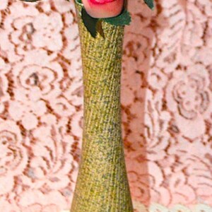 May include: A tall, slender, textured olive-green vase with a tapered neck, containing a bouquet of five artificial roses in shades of yellow, orange, and pink, each featuring green fabric leaves attached to the stems.
