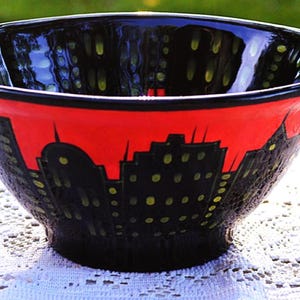 May include: A ceramic bowl featuring a black and red cityscape design. The exterior displays a silhouette of buildings in black against a vibrant red background, with small yellow dots representing illuminated windows throughout the structures.