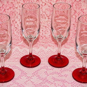 May include: A set of four clear glass champagne flutes, each featuring a slender stem and a vibrant, solid red circular base. The glasses have a tall, cylindrical bowl design with a smooth, transparent finish, standing upright in a symmetrical arrangement.
