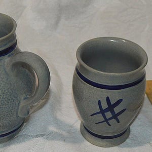 May include: A gray stoneware pitcher and a matching mug, both featuring a textured salt-glaze finish, dark blue horizontal bands, and a hand-painted blue cross-hatch pattern. The pitcher has a small handle and the embossed text 0.25.