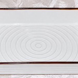 May include: A rectangular white ceramic baking dish with two side handles. The interior base features a raised concentric circle pattern. The top rim is finished with a thin brown border, and the exterior sides have horizontal ridges.
