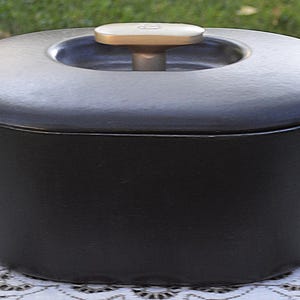 May include: A matte black oval Dutch oven with a matching lid and two side handles. The lid features a recessed center with a light-colored, T-shaped handle knob. The pot has a smooth, solid-colored exterior finish and a wide, deep cooking basin.