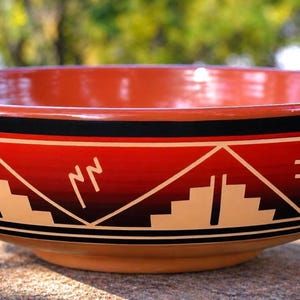 May include: A large, round ceramic bowl with a glossy finish. The exterior features a vibrant red base color decorated with black and cream-colored geometric patterns, including stepped pyramid shapes and linear symbols arranged in a repeating band.